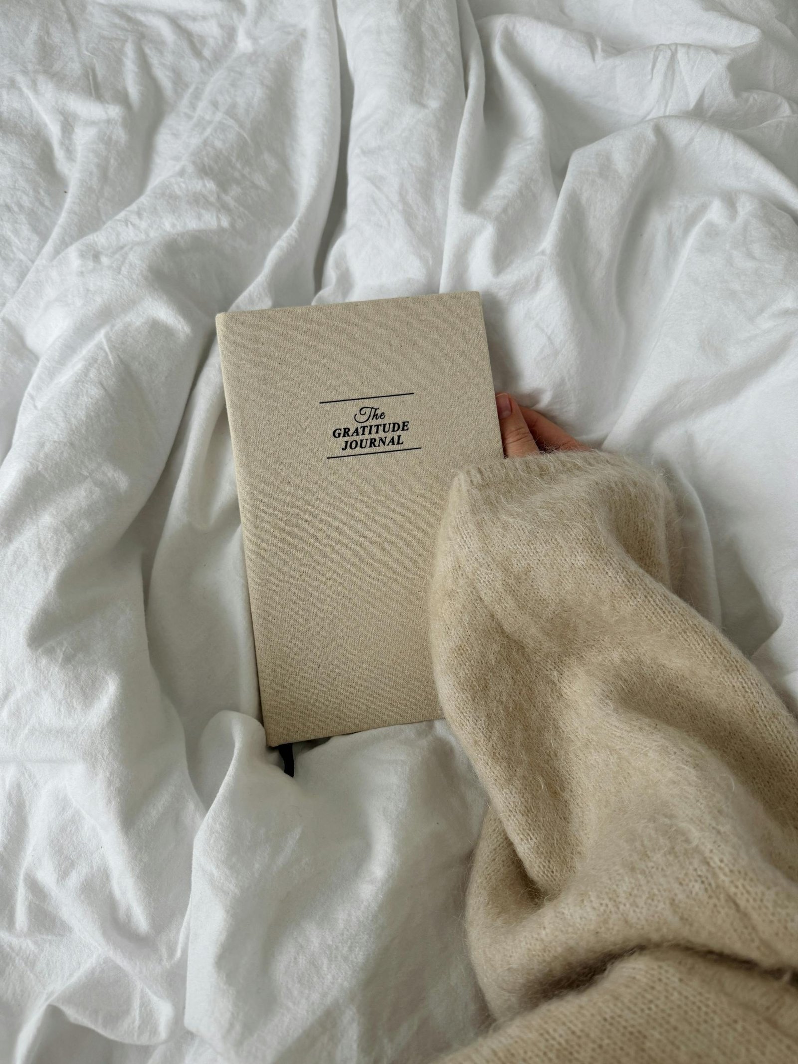 A close-up of a hand holding a gratitude journal on a soft bed, conveying warmth and reflection.